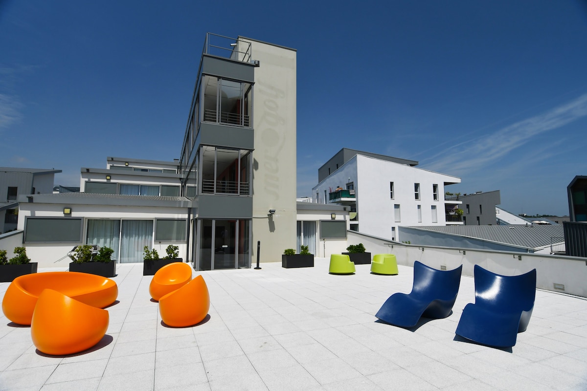A spacious outdoor terrace features modern lounge seating in vibrant orange and deep blue colors. The area is bathed in sunlight, providing a bright and open atmosphere. Surrounding planters add greenery to the minimalist design of the terrace and are complemented by the contemporary architecture of the building.