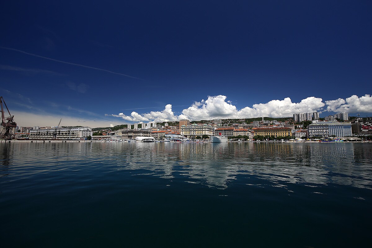 A panoramic view of Rijeka is presented, with the city skyline reflecting on the calm water. Buildings in a variety of colors line the waterfront, set against a backdrop of a bright blue sky dotted with white clouds.