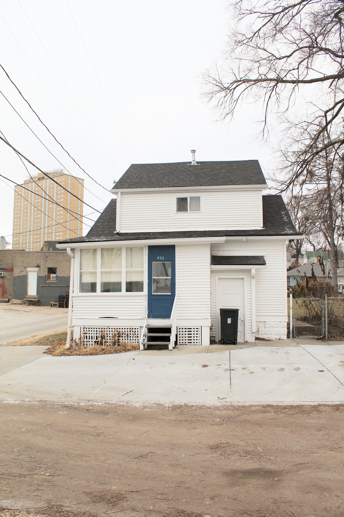 The exterior of the house is displayed, showcasing a two-story structure with white siding and a dark roof. A blue front door is visible, along with a small porch area. A concrete driveway is present, and utility connections are noted beside the home.