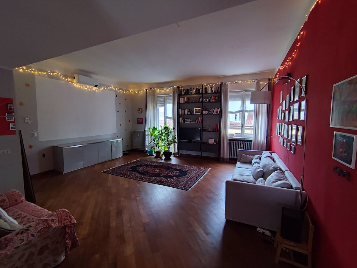 A spacious living area features wooden flooring and a blend of red and neutral walls. Soft seating is arranged around a traditional rug, with a tall bookshelf displaying various books. Potted plants add greenery, while soft lighting creates a warm ambiance.