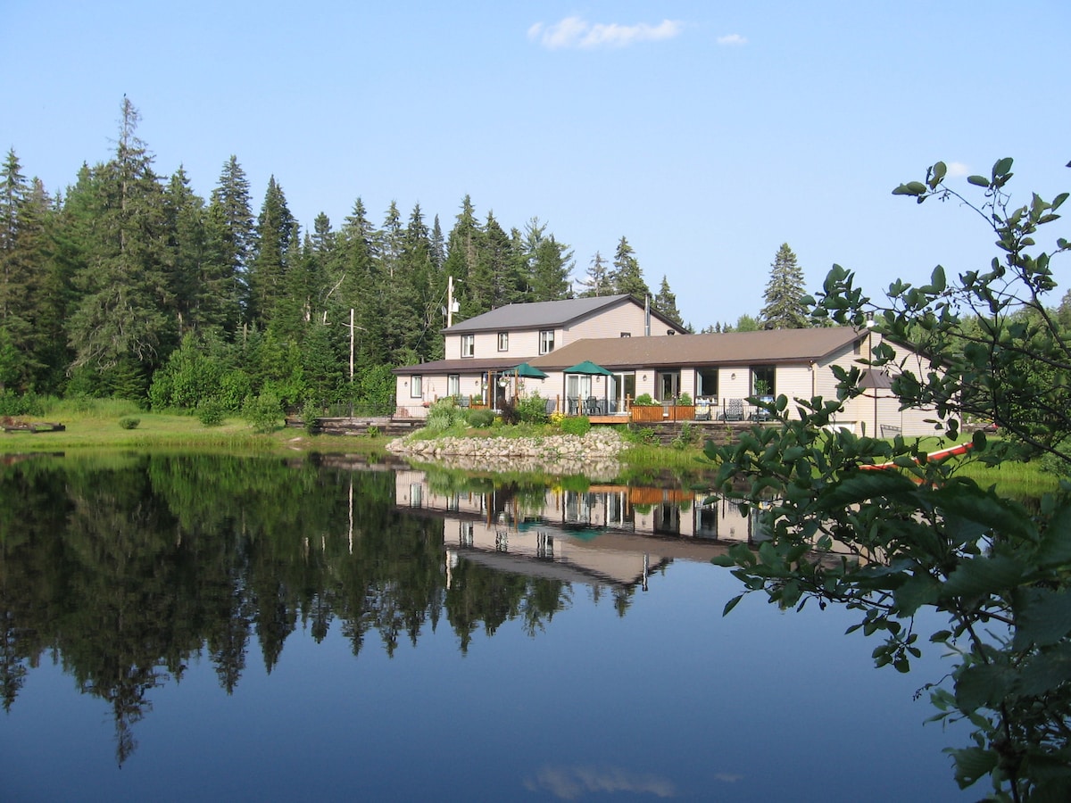 A lakeside lodge is reflected in the serene waters of Lac Ouimet, surrounded by lush greenery and towering trees. The structure features a spacious patio with seating and umbrellas, inviting relaxation amidst the natural landscape.