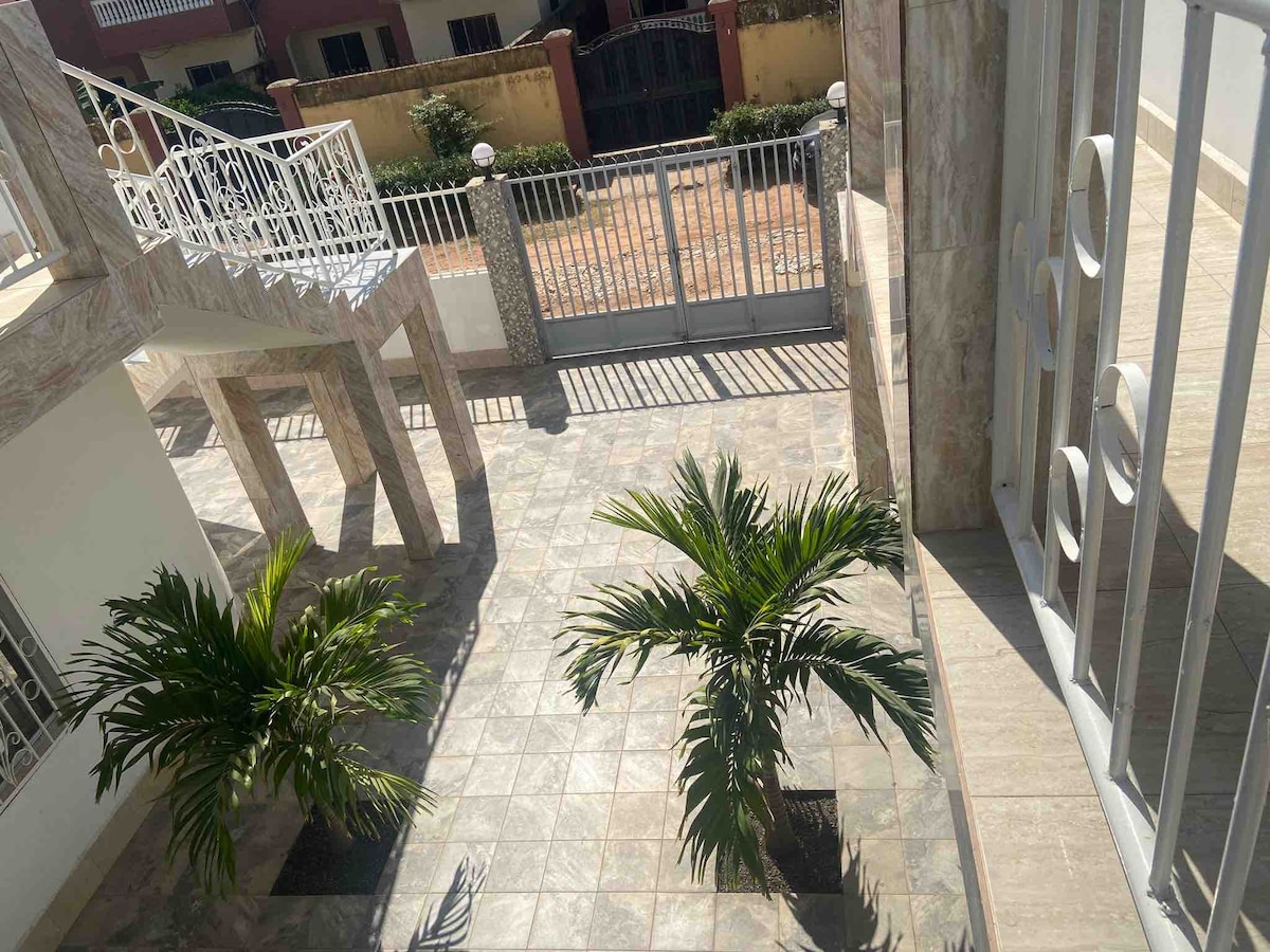 The image displays a courtyard area featuring two potted palm trees surrounded by tiled flooring. The surroundings are characterized by a gated entrance and a staircase leading to an upper level, with sunlight casting shadows on the ground.