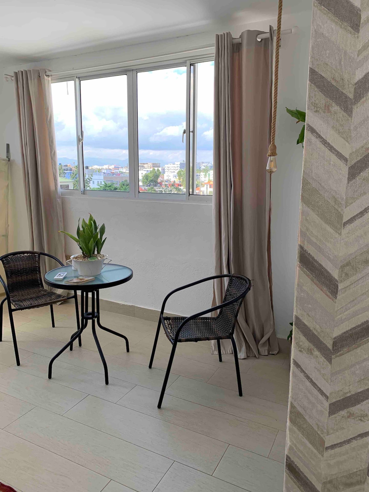 A small dining area is seen with two black chairs positioned around a circular table. Light-colored curtains frame a large window that offers views of the surrounding cityscape and greenery. A potted plant sits on the table, enhancing the natural ambiance.