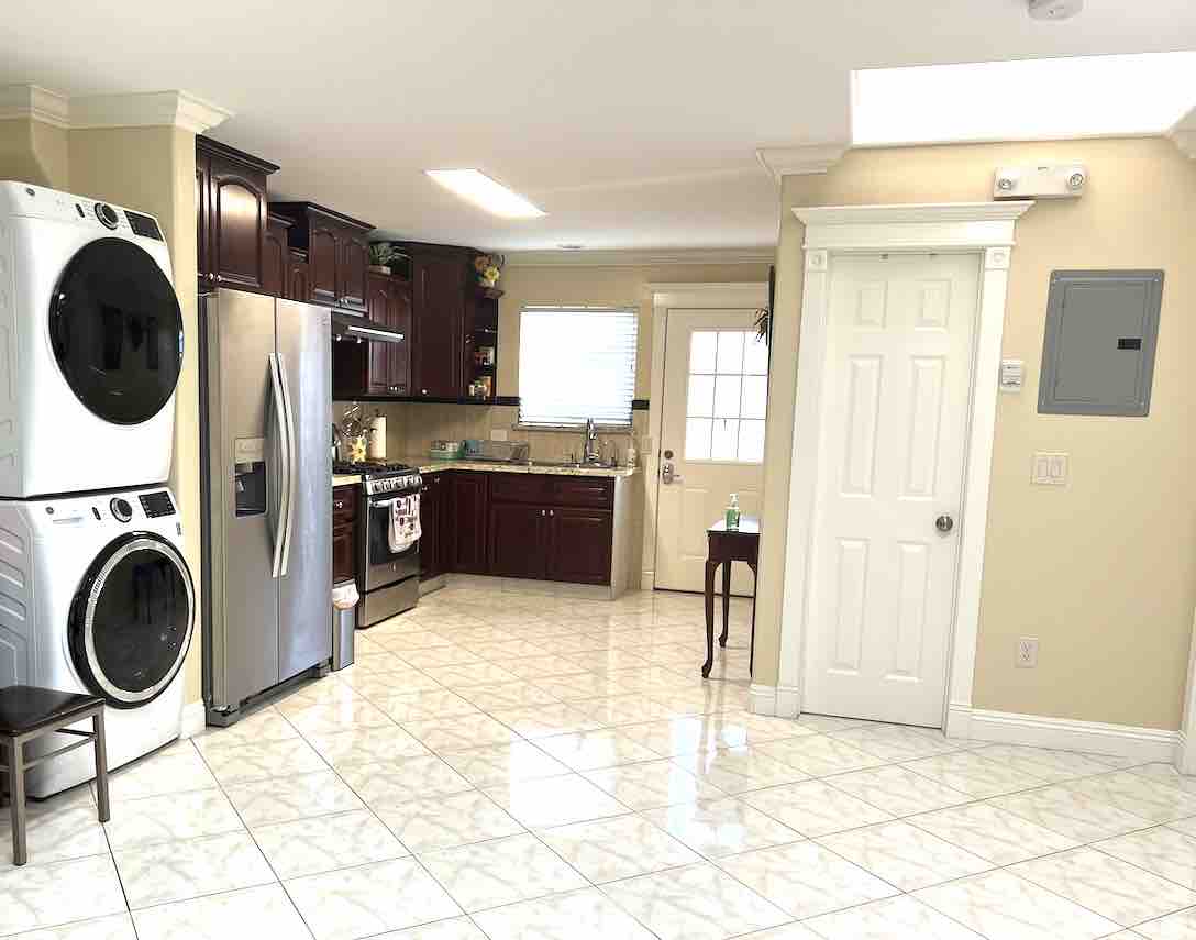 The kitchen area showcases dark wood cabinetry, stainless steel appliances, and a spacious layout. A washer and dryer are positioned to the left, with a door leading to the exterior at the back. Natural light illuminates the tiled floor, enhancing the room's overall brightness.