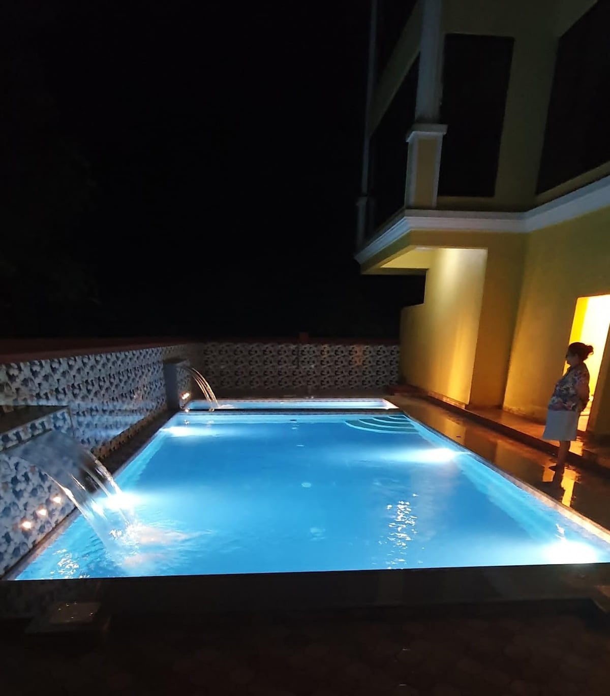 A tranquil swimming pool is illuminated in soft blue light, enhancing the serene evening setting. Water features create gentle splashes, while the surrounding walls add privacy. A warm glow emanates from the nearby building, providing a welcoming backdrop for relaxation.