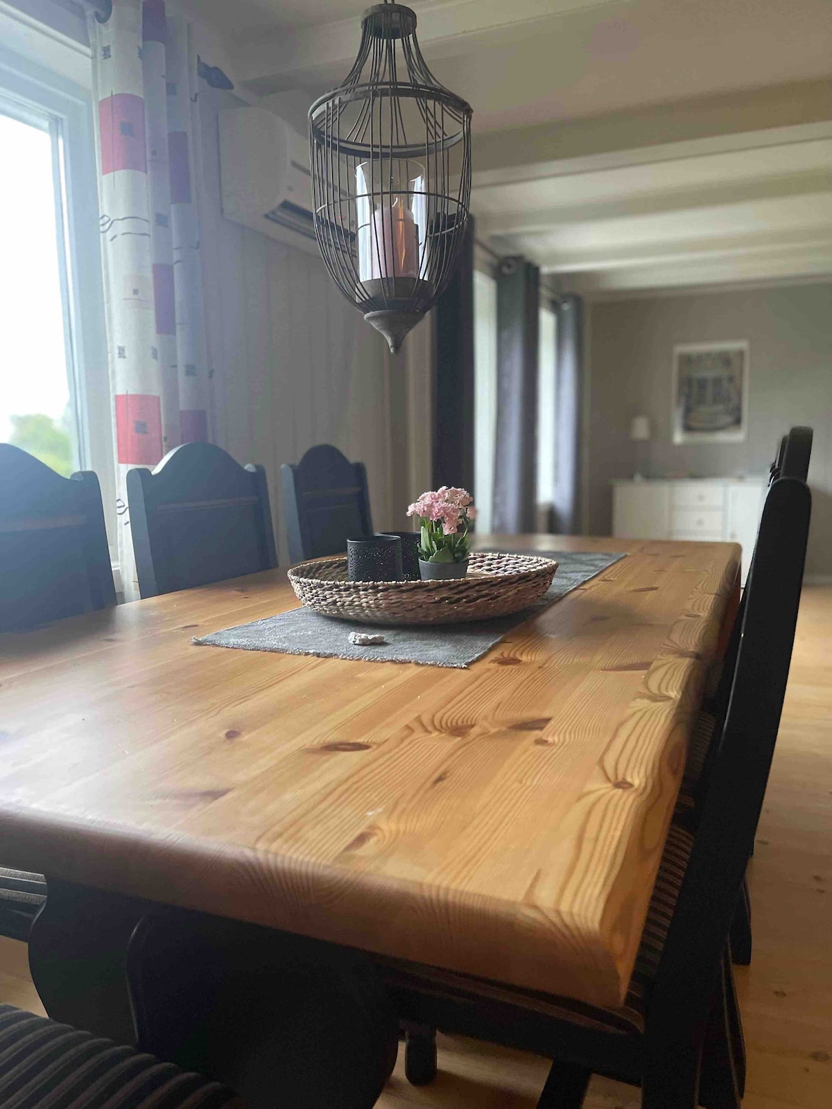 A spacious dining table made of natural wood is set with a decorative tray containing a small potted flower. Surrounding the table are several black wooden chairs. A pendant light hangs above, illuminating the area through a nearby window with sheer curtains.