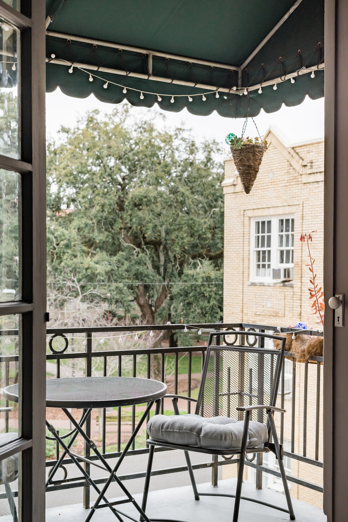 A small balcony features a round bistro table and a single chair, providing a cozy space. Hanging greenery adds a natural touch, while views of lush trees and the surrounding historic architecture are visible. The balcony is partially sheltered with a green awning.