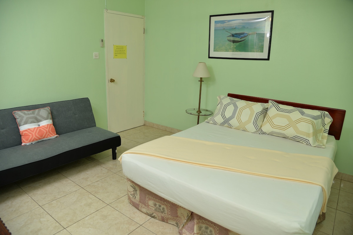 A ground floor room features a queen bed with a light-colored bedspread, complemented by decorative pillows. A futon sits beside the bed, and a small table with a lamp stands adjacent. A framed photograph of a boat decorates the green wall.