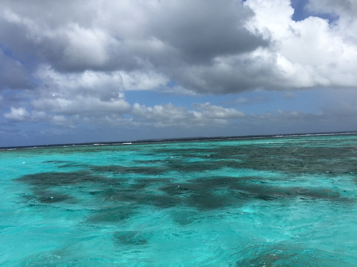 The image captures a panoramic view of the lagoon, featuring vibrant turquoise waters layered with darker patches. Soft clouds drift across a bright sky, creating a serene atmosphere. The horizon extends into the distance where the sea meets the sky.