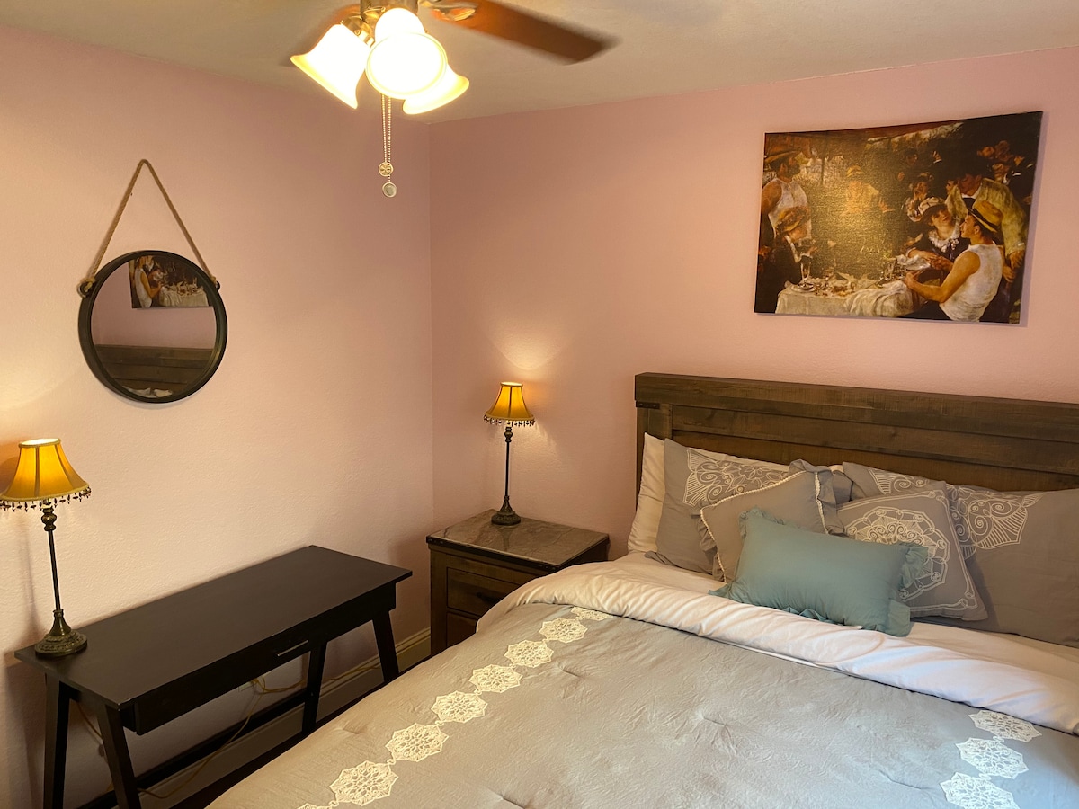 A cozy room features a spacious bed with light bedding, a decorative pillow, and framed artwork above. A vanity desk with a simple design and a lamp are positioned next to the bed, with another lamp on the bedside table. The walls are painted a soft pink.