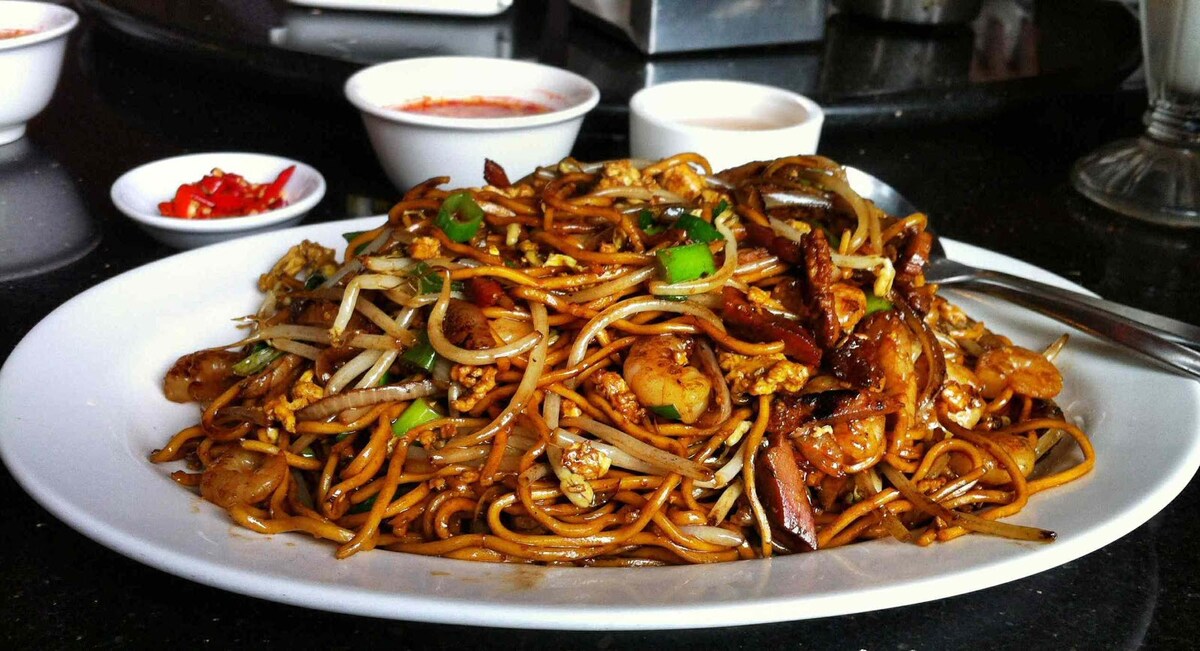 A plate of stir-fried noodles is presented, garnished with green onions and accompanied by small bowls of sauces. The dish includes various seafood and vegetables, showcasing a colorful and appetizing arrangement.