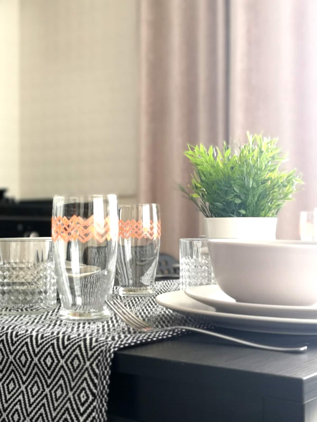 The dining table is set with various glassware, featuring decorative patterns. A small plant in a white pot adds a touch of greenery. Plates and bowls are neatly arranged, complemented by a patterned table runner that enhances the overall presentation.