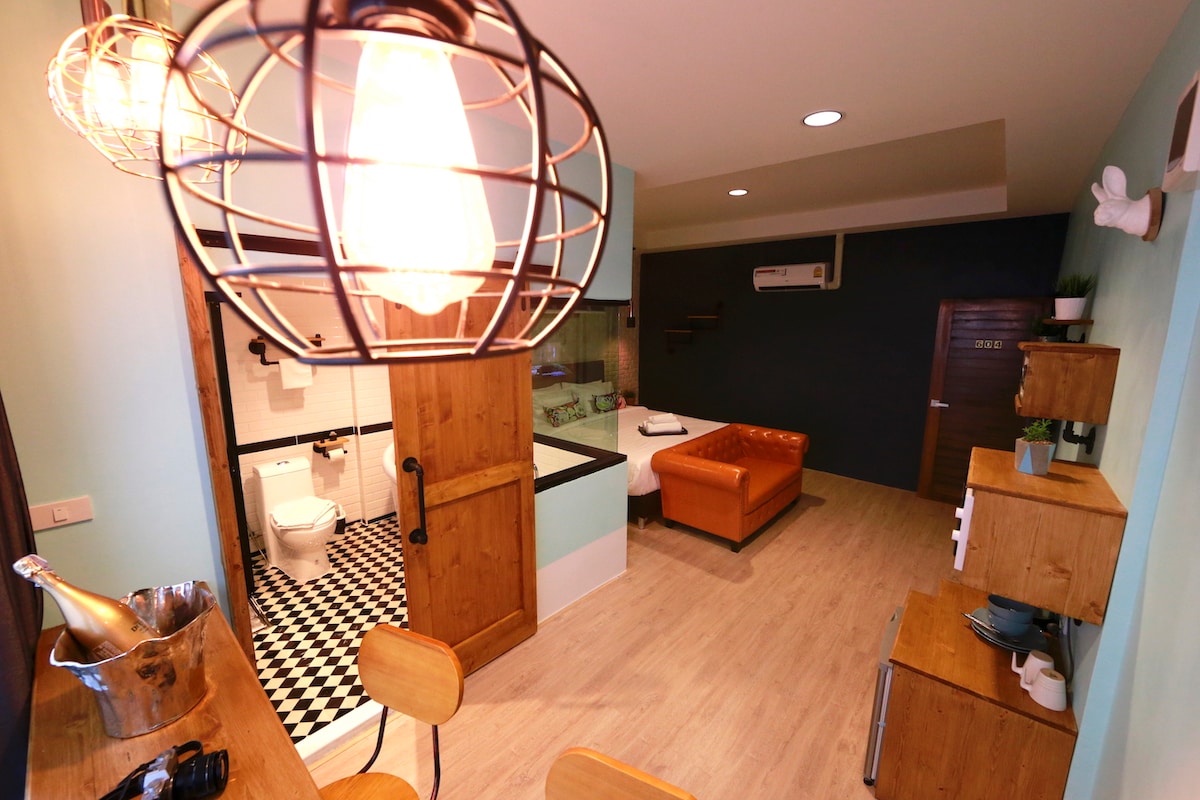 A modern room is showcased with wooden accents and a spacious layout. A cozy sofa in the corner complements a well-equipped kitchenette. The dining area features two stools, while a stylish light fixture hangs above. The bathroom is visible through a partially open doorway, with a classic black and white tile design.