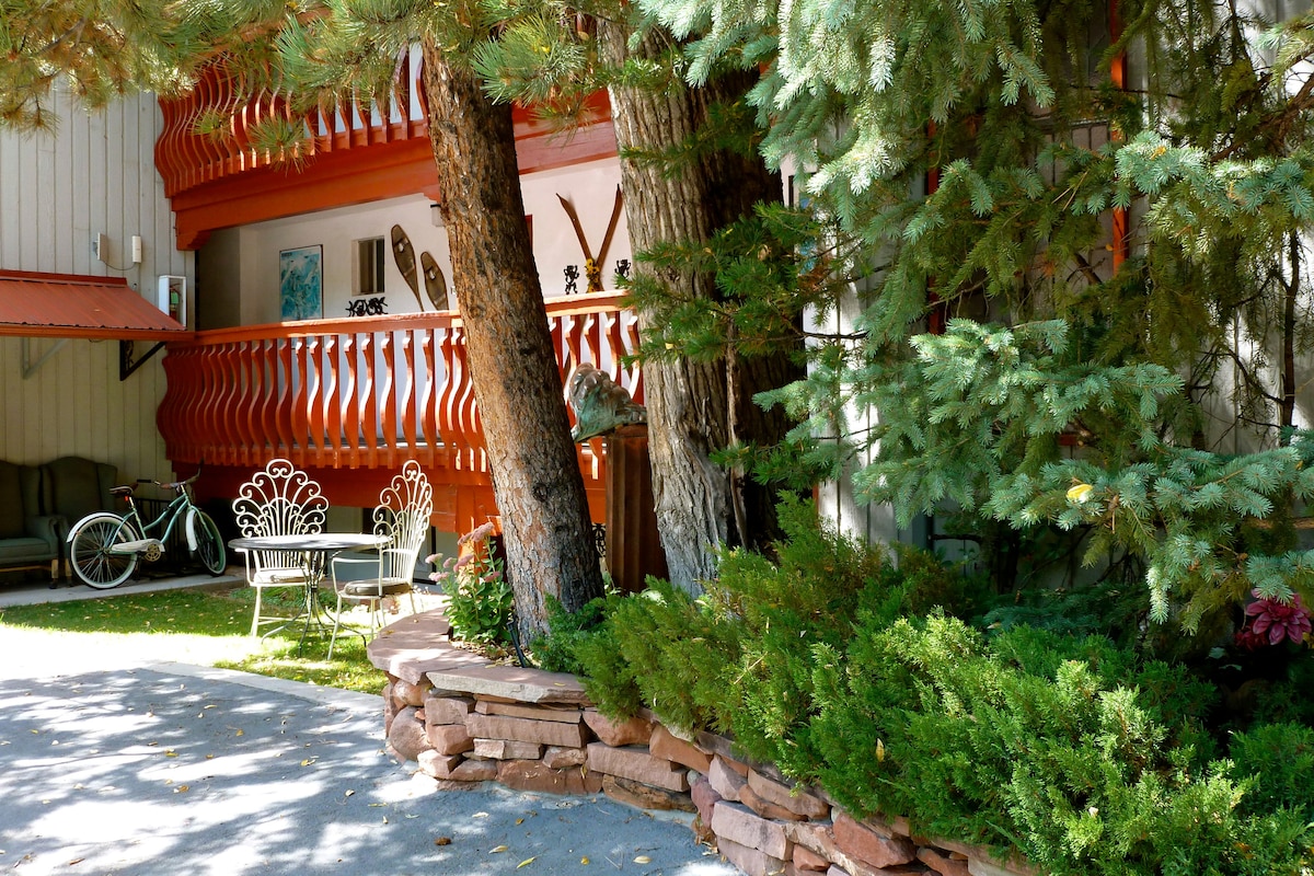 A peaceful outdoor area features a stone border with lush greenery and a small seating arrangement. Two vintage bicycles can be seen nearby, while wooden skis hang decoratively on the balcony. The space is shaded by mature trees, providing a serene atmosphere.
