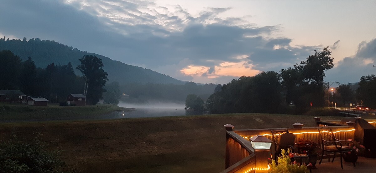 A serene view of rolling hills and a foggy river during twilight, framed by trees. The outdoor deck is illuminated with soft lighting, creating a calming atmosphere. Gentle clouds scatter across the sky, hinting at the day's end, while the landscape blends into soft silhouettes.