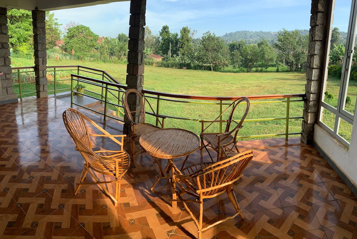 A spacious porch area is highlighted with a circular wooden table and four rattan chairs, providing a comfortable seating arrangement. Lush greenery and rolling hills are visible in the background, with large windows allowing natural light to fill the space.