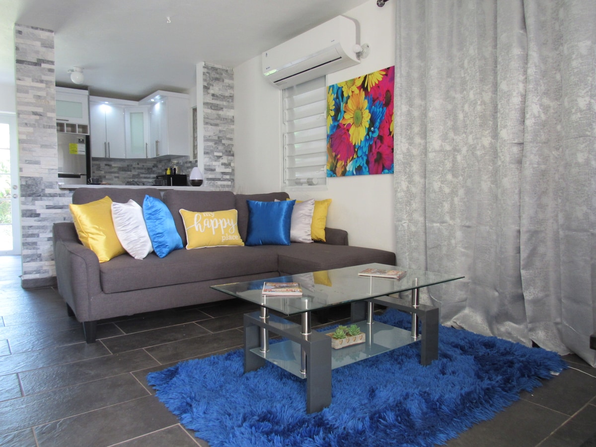 A modern living area features a plush gray sofa adorned with colorful pillows. A glass coffee table sits atop a vibrant blue rug, while a piece of abstract wall art adds a splash of color. Natural light enters through a nearby window, enhancing the space.