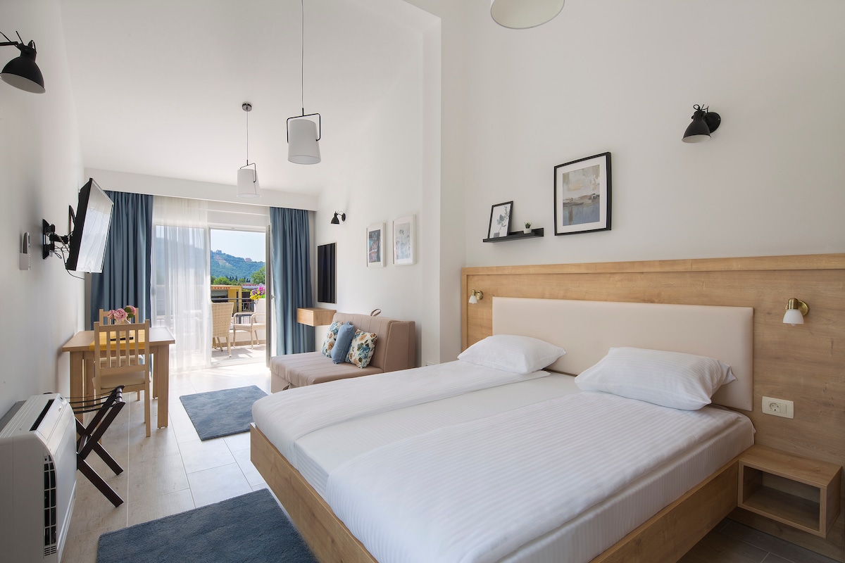 A bright studio apartment features a spacious bed and a comfortable sofa-bed. Natural light floods the room through large glass doors leading to the terrace. A wooden dining table is positioned nearby, while decorative wall art and lighting enhance the modern design.