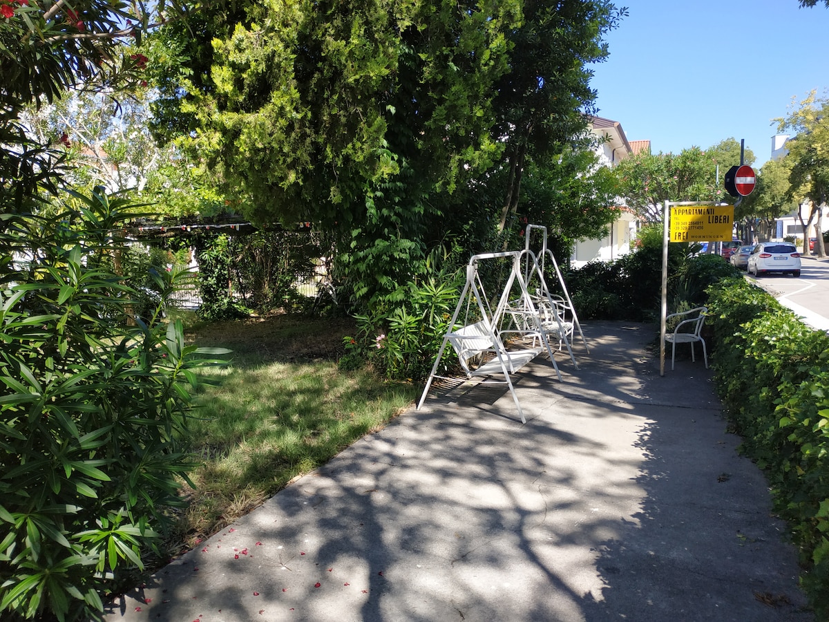 A shaded walkway lined with greenery features two white swing benches set on a concrete path. The surrounding plants create a natural barrier, while sunlight filters through leaves, casting dappled shadows on the ground.
