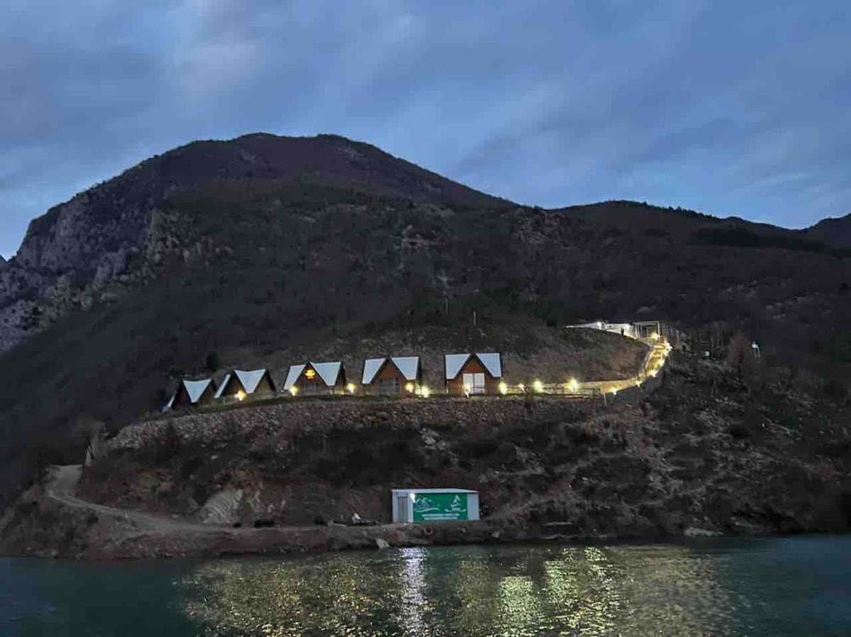 A cluster of triangular-roofed cabins is positioned on a hillside, softly illuminated against the twilight sky. The landscape features rocky terrain leading to a calm body of water, reflecting the gentle glow of the cabin lights.
