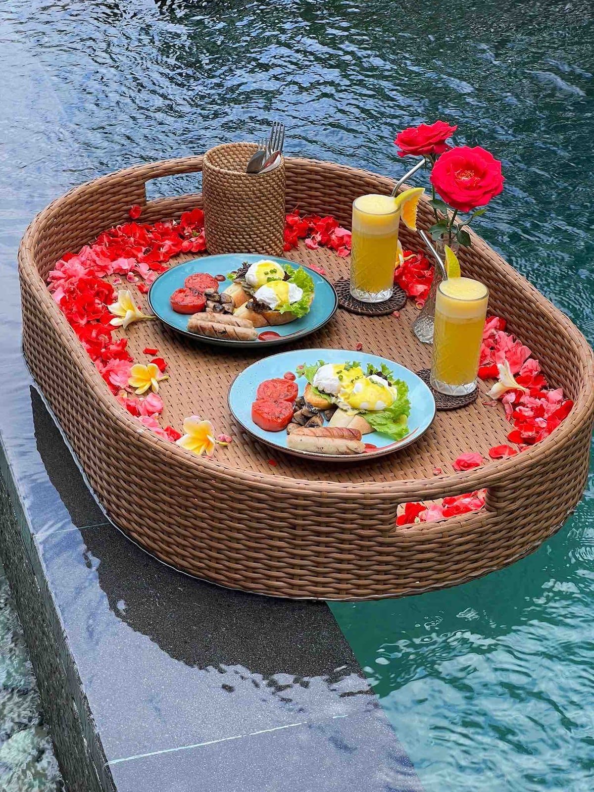A woven tray is placed on the edge of an outdoor swimming pool, featuring two plates of breakfast and two glasses of orange juice. The tray is adorned with vibrant red flower petals and tropical blossoms, enhancing the dining experience in this serene setting.