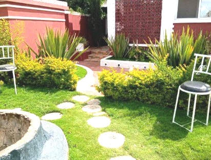 A serene outdoor space features a winding stone pathway leading through lush greenery and low hedges. Two white chairs are positioned nearby, inviting relaxation. The surrounding plants create a tranquil atmosphere, complemented by a small circular feature in the foreground.