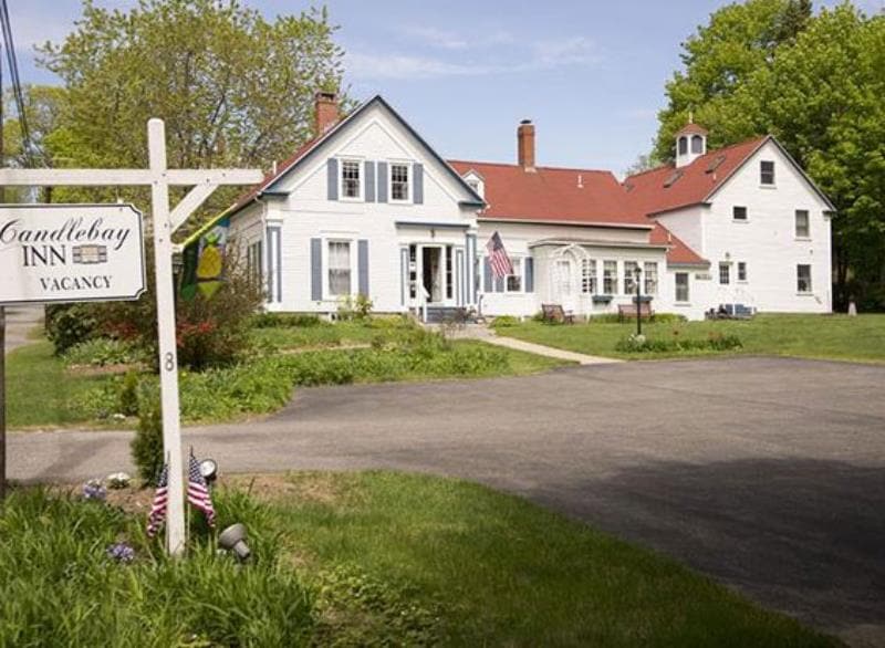A historic inn is depicted with a white exterior and blue accents, complemented by a welcoming front porch. The manicured lawn is lined with flowering plants and flags. A paved driveway leads to the entry, suggesting ease of access and parking for guests.