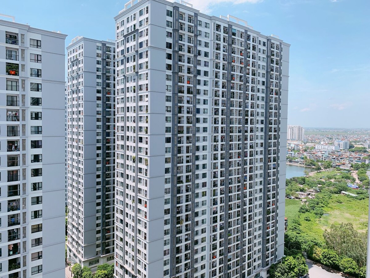 A view of modern high-rise apartment buildings showcases a contemporary architectural style. Lush greenery is visible at the base, along with a river flowing nearby. The scene is set under a clear blue sky, emphasizing the urban landscape.