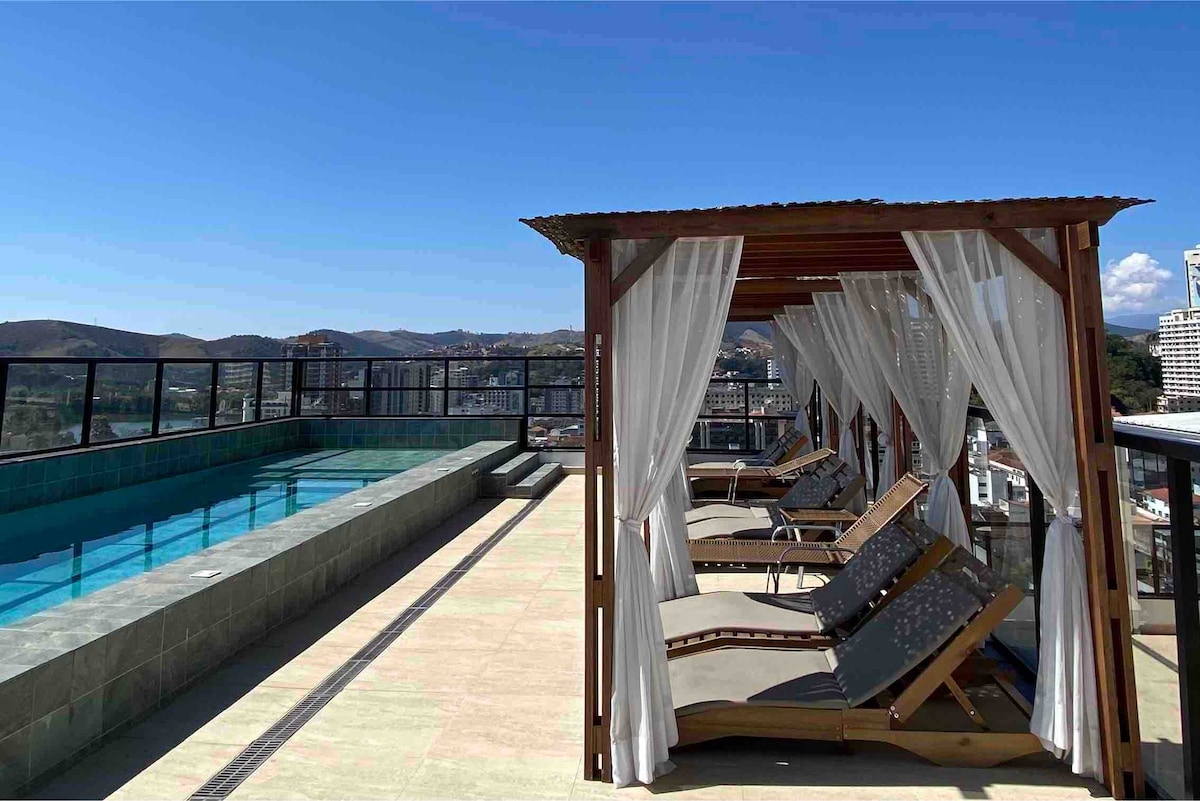 An outdoor rooftop area features a pool alongside a shaded seating structure with draped curtains. Comfortable lounge chairs are arranged on the deck, providing views of the surrounding city and mountains under a clear blue sky.