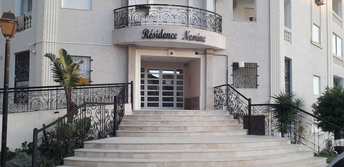 The exterior of the building features a grand entrance with a wide staircase leading up to double glass doors. Ornate metal railings frame the stairs, complemented by landscaped greenery. The name 'Résidence Ménime' is prominently displayed above the entrance.