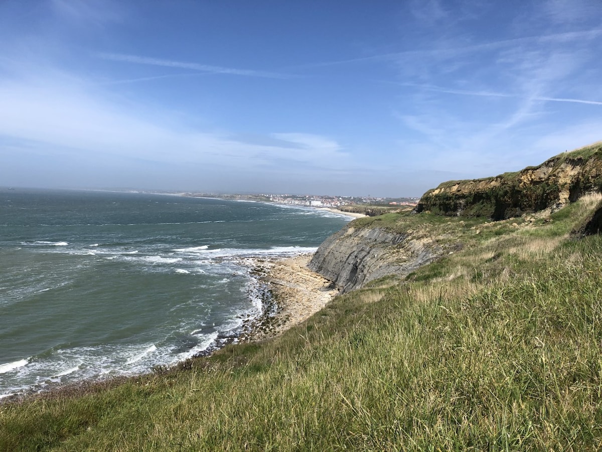 A coastal landscape is presented, with rolling green hills leading down to rocky shorelines. Gentle waves break against the rocks, while a distant shoreline features a small town under a clear blue sky with occasional wispy clouds.
