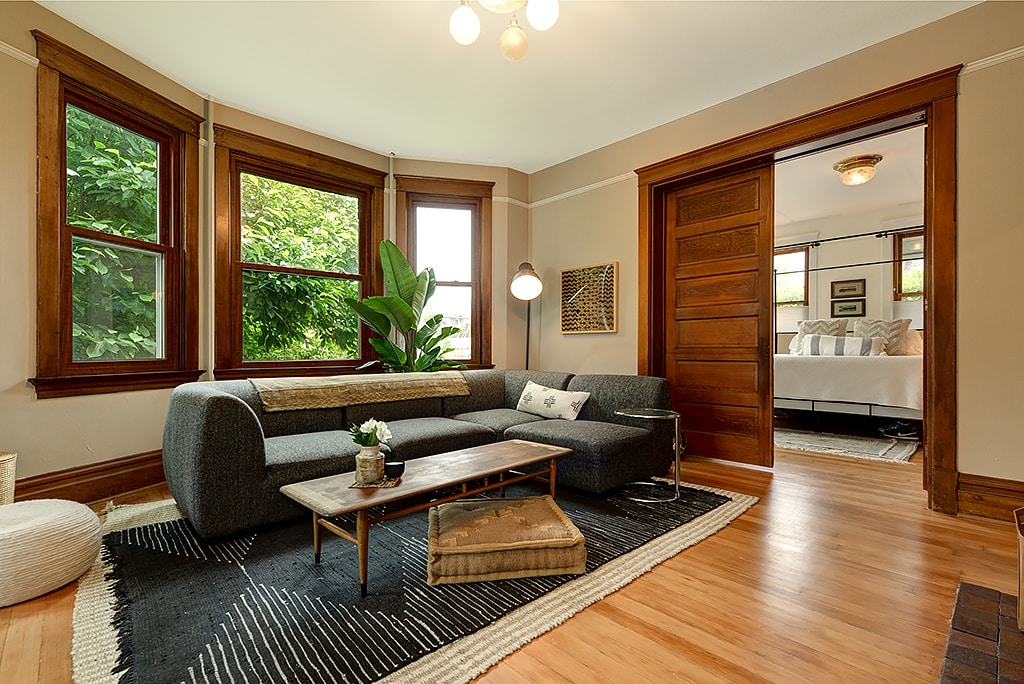 A cozy living area features a modern sectional sofa in gray, accompanied by a coffee table and a standing lamp. Large windows allow natural light to fill the room, with greenery visible outside. The warm wood floors and decorative area rug add a welcoming touch.