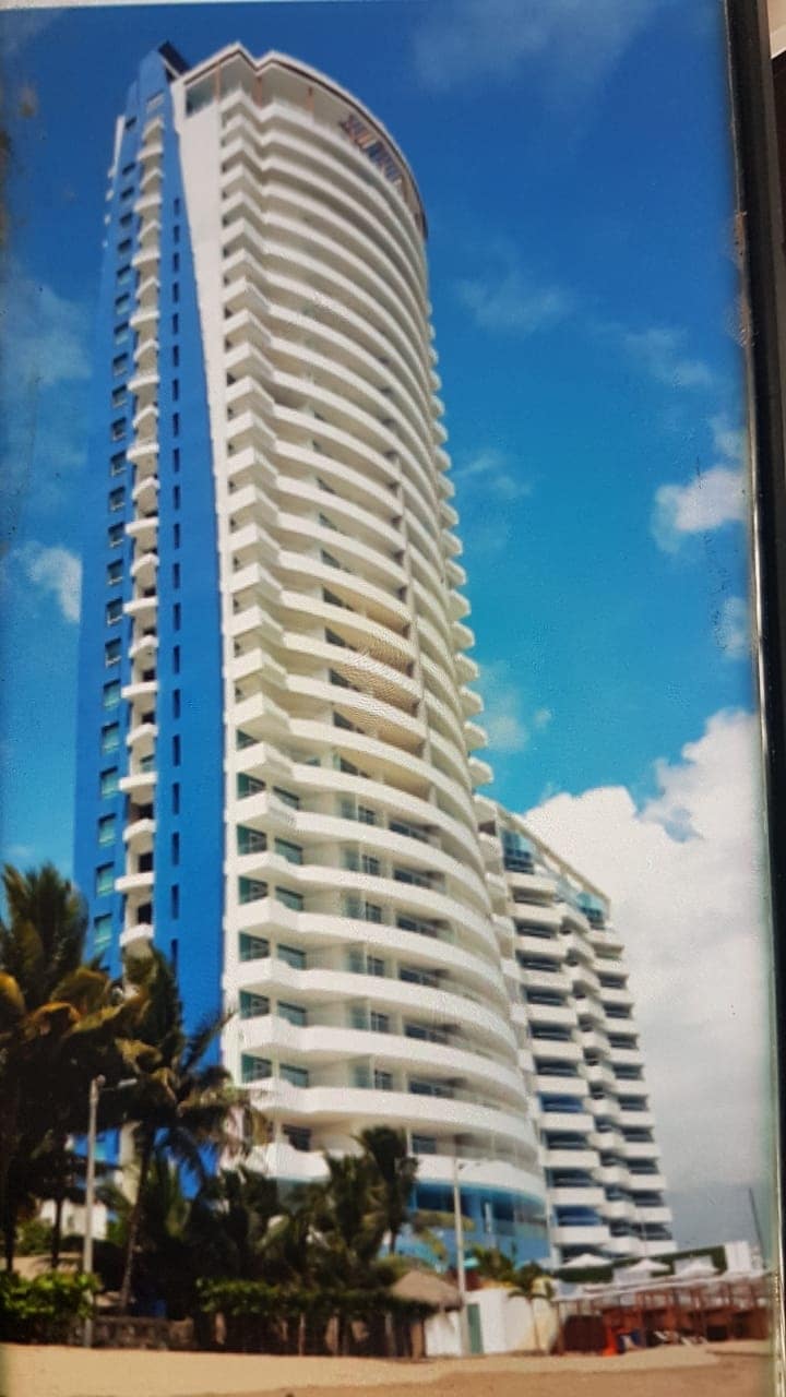 A modern high-rise building features a sleek, curved facade with a mix of white and blue panels. Multiple balconies are visible on each floor, providing an open and airy architectural style. The sky in the background is clear with scattered clouds.