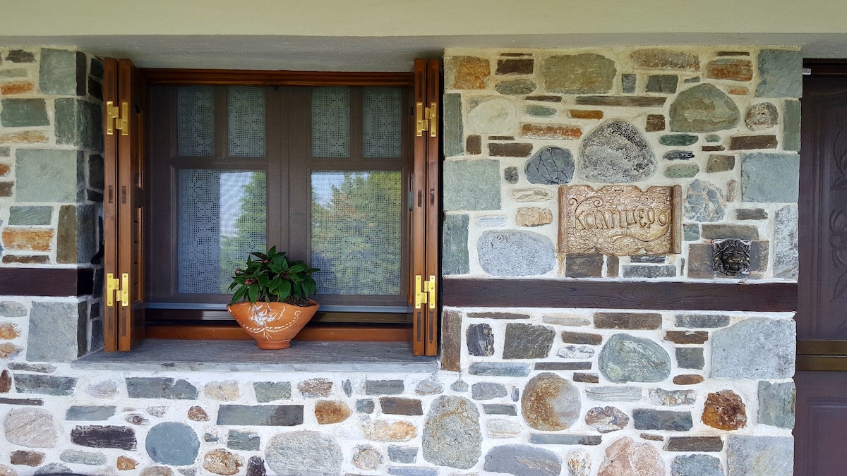 A stone wall features a window with wooden shutters, opened to reveal a small potted plant on the windowsill. A decorative stone plaque with carved letters is positioned next to the window, contributing to the traditional local architecture.