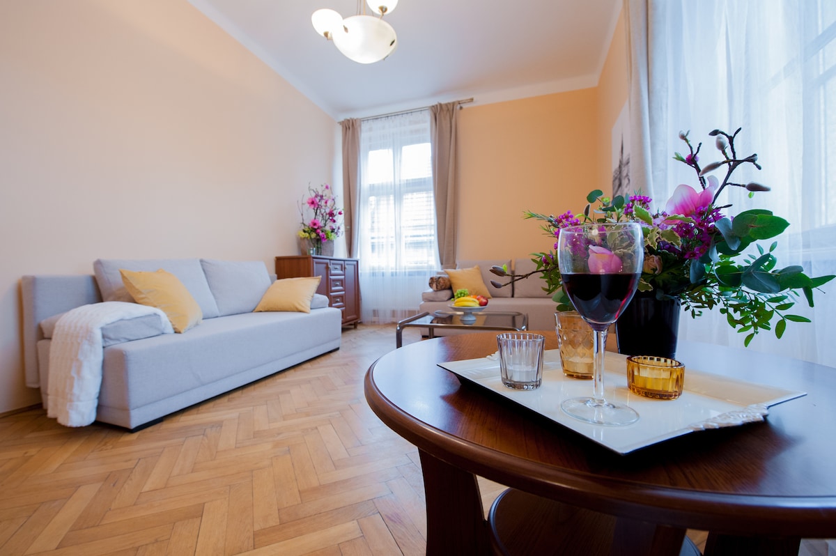 A bright living area is highlighted by a comfortable gray sofa adorned with yellow cushions. A wooden table displays a decorative arrangement of flowers and glasses, complemented by a bowl of fresh fruit. Large windows allow natural light to fill the room.