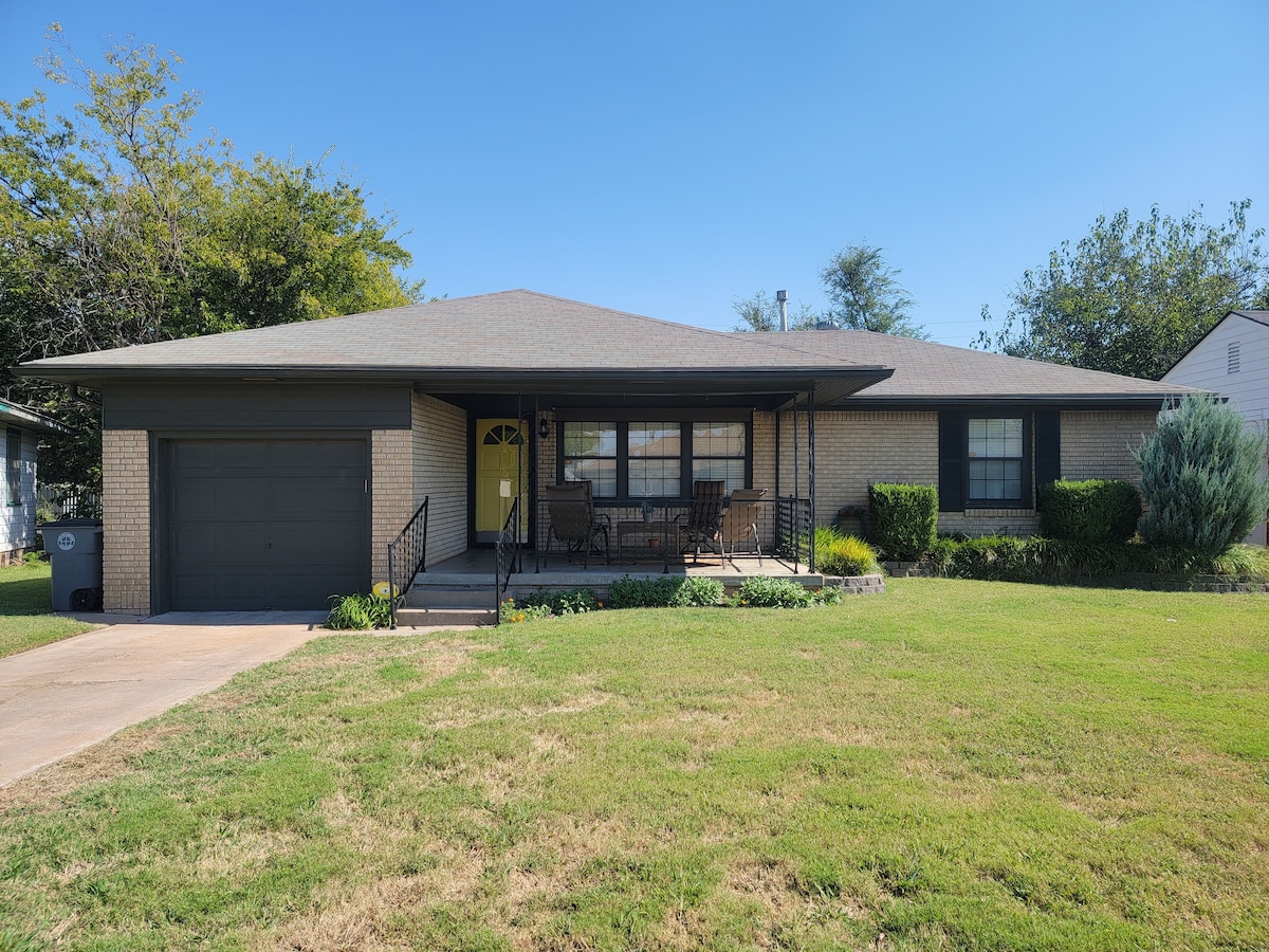 A modern home is presented with a welcoming front porch featuring seating and surrounded by a well-maintained lawn. The garage is visible, and shrubs add greenery to the landscape. The structure is complemented by a clear blue sky above.