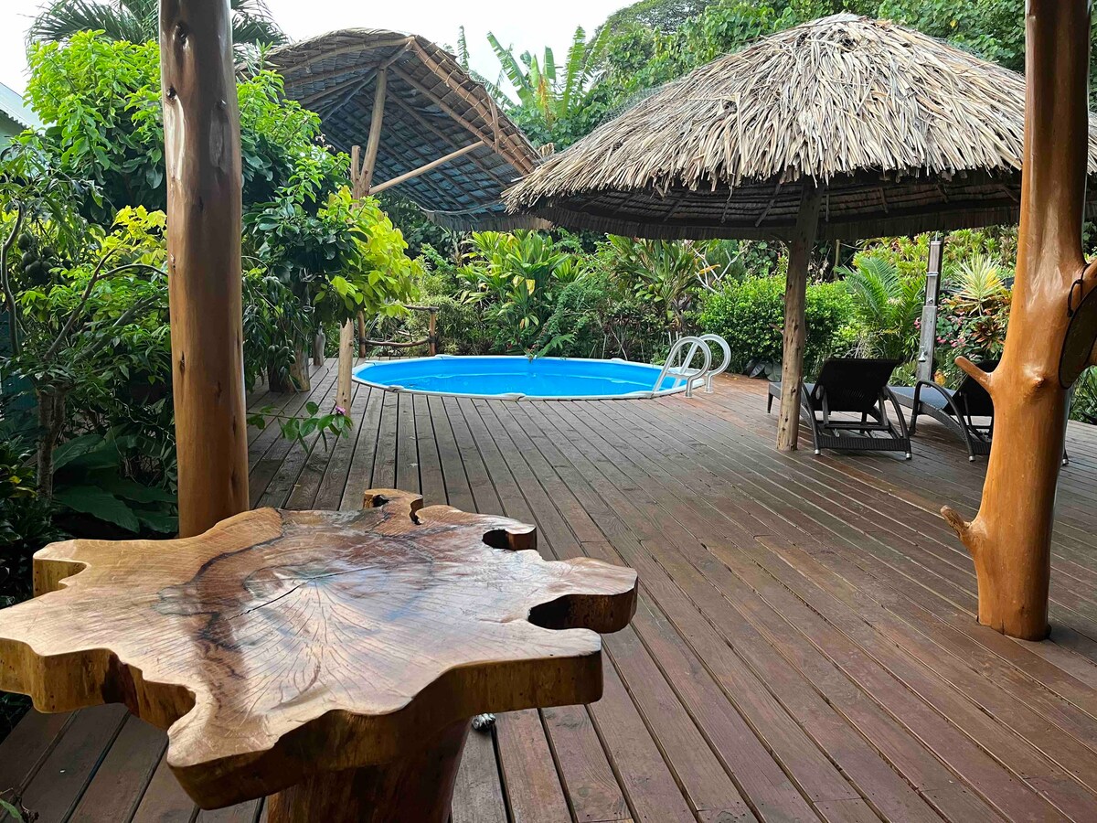 A wooden deck surrounds a blue swimming pool, bordered by lush greenery. Two thatched umbrellas provide shade over lounge chairs, offering a cozy spot for relaxation. The wooden table in the foreground adds a natural touch to the inviting outdoor space.