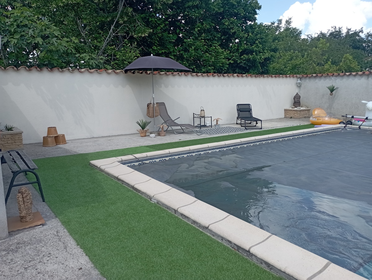 A secure swimming pool area features a large pool covered with a safety tarp. Lounge chairs and an umbrella provide shaded seating, accompanied by potted plants. An artificial grass border runs alongside the pool, enhancing the overall relaxation space.