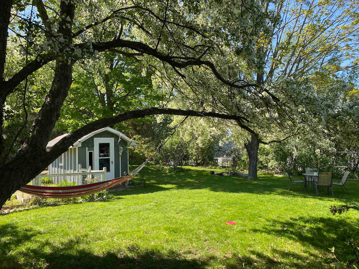 A shaded backyard is showcased, featuring a hammock draped between two trees and a lush green lawn. A small shed stands in the background, and a patio set is visible to the right, surrounded by flowering trees that provide a tranquil setting.