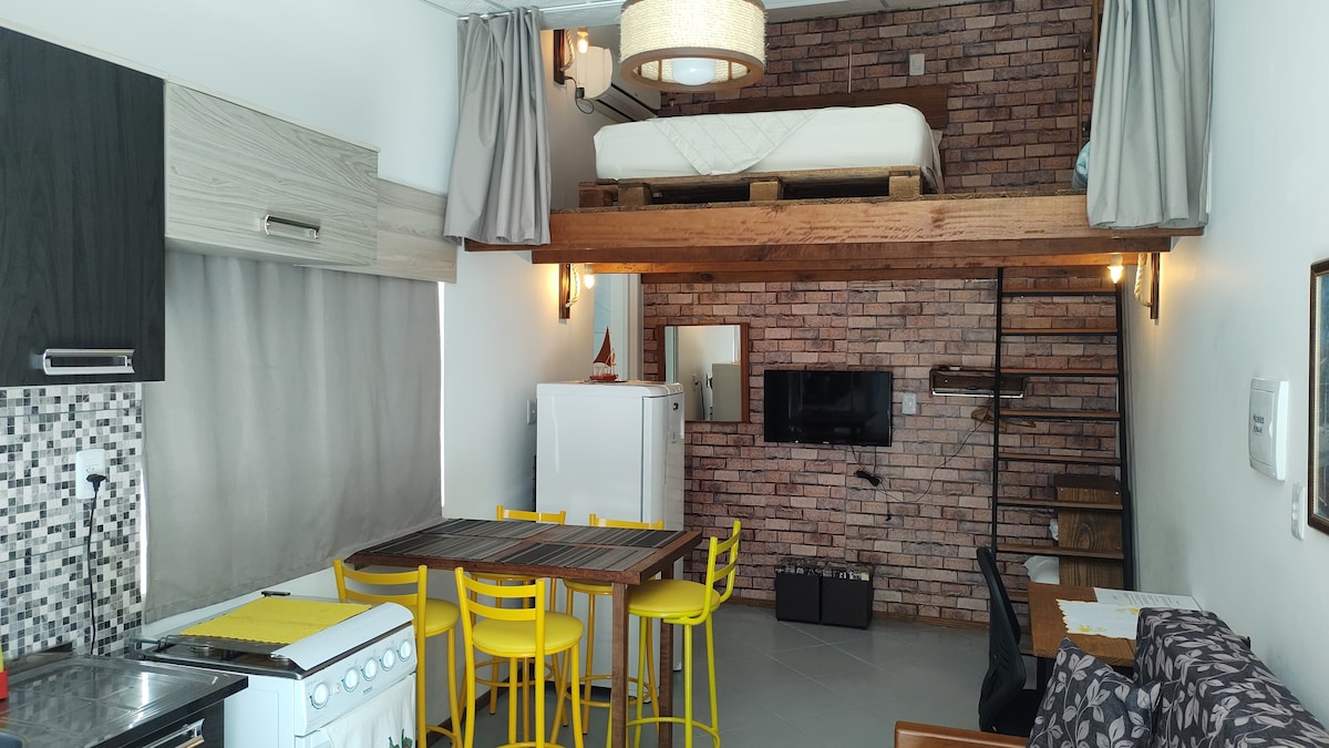 An open loft space features a cozy sleeping area on an upper mezzanine. A wooden staircase provides access to the bed above. Below, a dining table with yellow chairs is positioned near modern kitchen appliances, while a comfortable seating area is complemented by exposed brick walls.