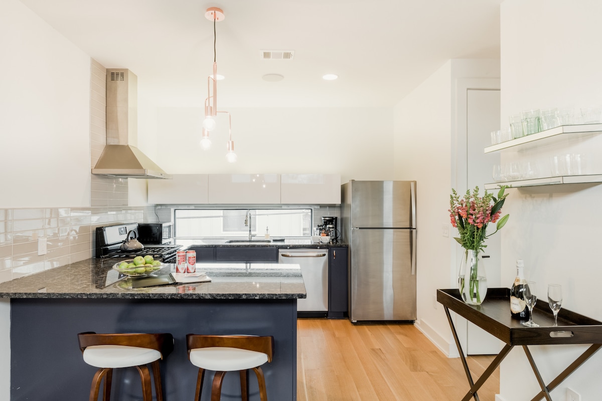 A modern kitchen is equipped with sleek granite countertops, stainless steel appliances, and a gas stove. Bar seating is available at the island, with subtle pendant lighting overhead. Fresh flowers add a touch of color, while glassware is neatly arranged on an adjacent shelf.