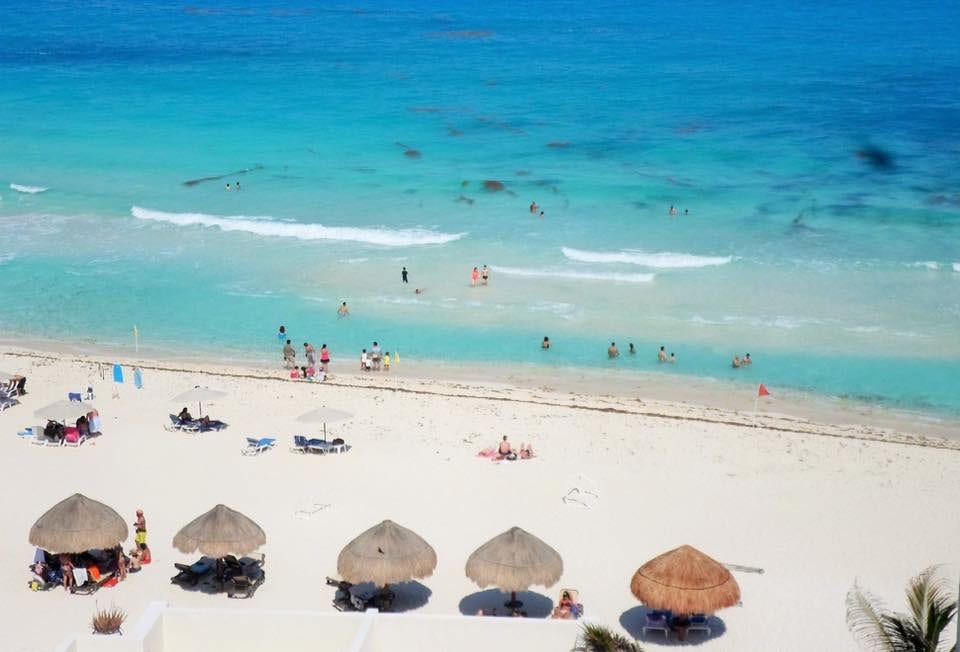 The image displays a sandy beach with clear turquoise waters and gentle waves. Several sun loungers and palapas are visible along the beach, where people can be seen enjoying the sun and water activities. The serene ocean extends into the horizon.