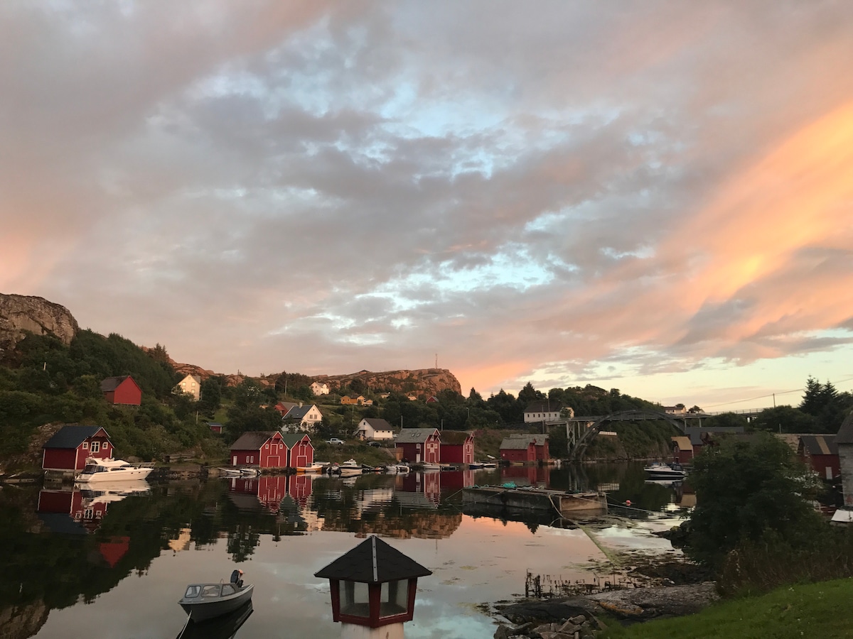 A serene landscape shows colorful fishing huts reflecting in calm waters during sunset. The sky is painted in soft pastels, and boats are visible on the water, surrounded by lush greenery and rocky hills in the background.