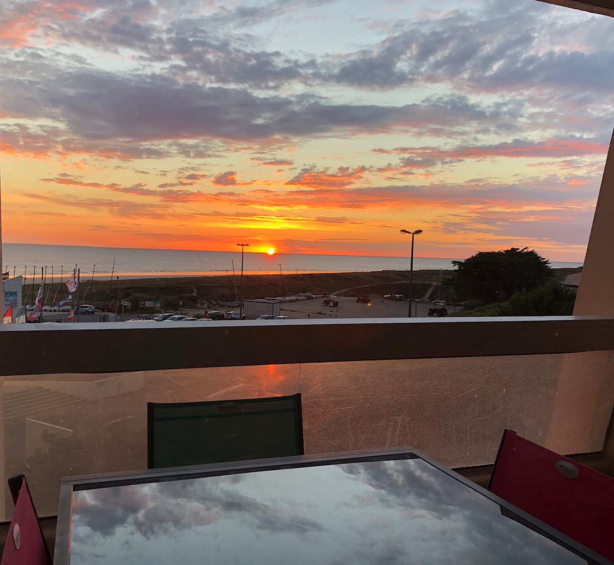 A seaside view is captured from the balcony, showcasing a vibrant sunset over the horizon. Colors of orange and pink blend in the sky, reflected subtly on the table surface. The setting evokes a sense of tranquility by the coast.