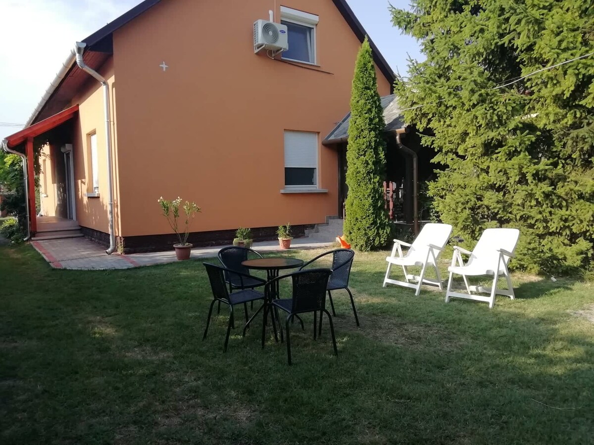 A spacious outdoor area features a manicured lawn with two white lounge chairs and a round black table surrounded by four chairs. Lush green trees provide shade, and the house's orange exterior is complemented by a small patio area.