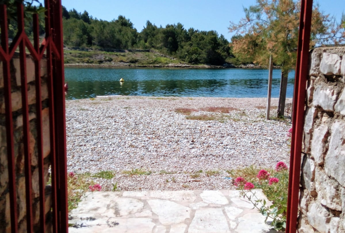 A view of the serene gravel beach and calm blue waters is framed by an open gate. Lush greenery can be seen in the background, enhancing the natural setting. The pathway consists of stone slabs, leading toward the water's edge dotted with vibrant flowers nearby.