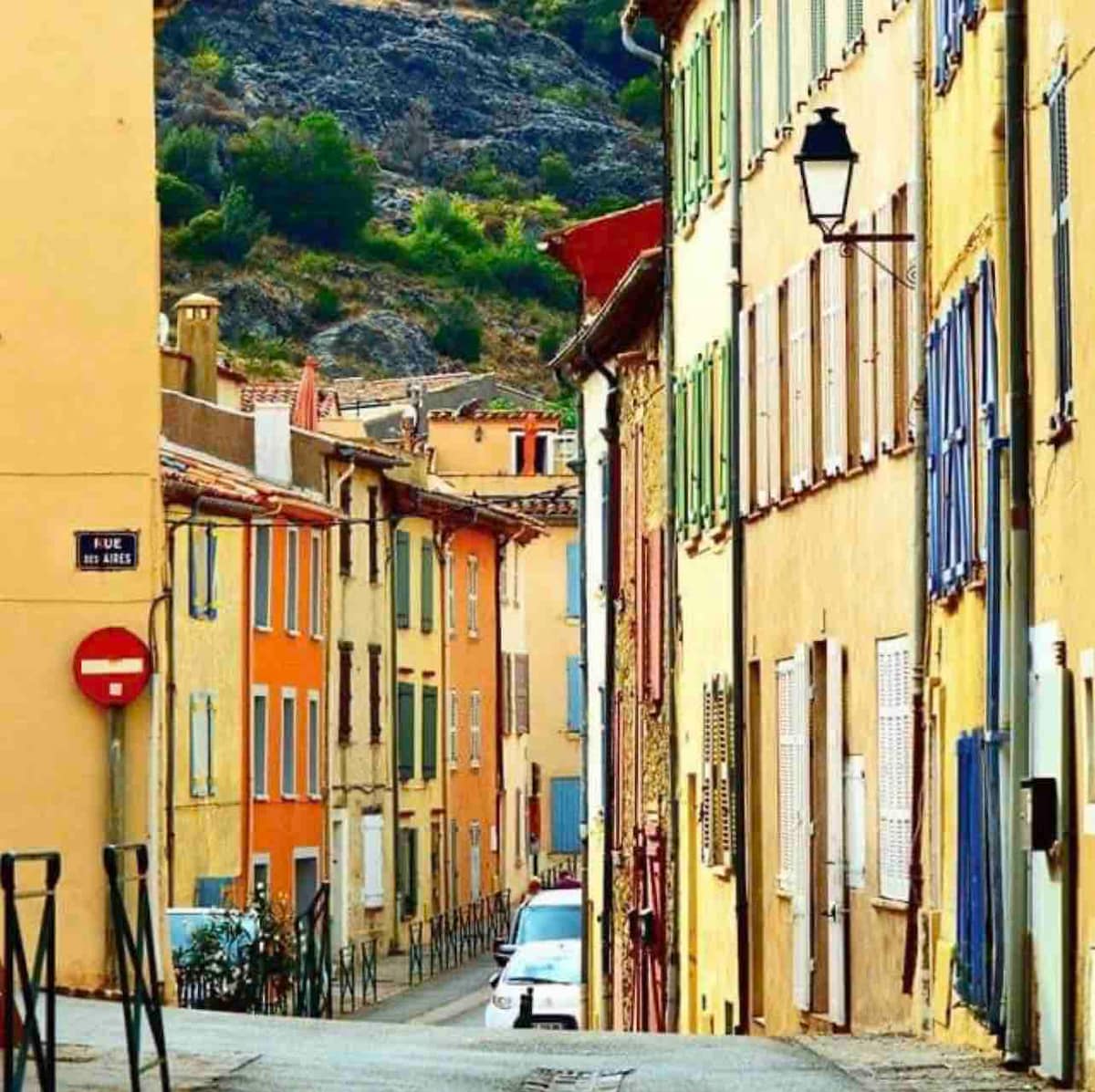 A narrow street in a village is lined with traditionally colored buildings featuring wooden shutters and textured facades. The street is observed descending gently, with greenery in the background and a few parked vehicles visible. Street signs and light fixtures add to the local charm.