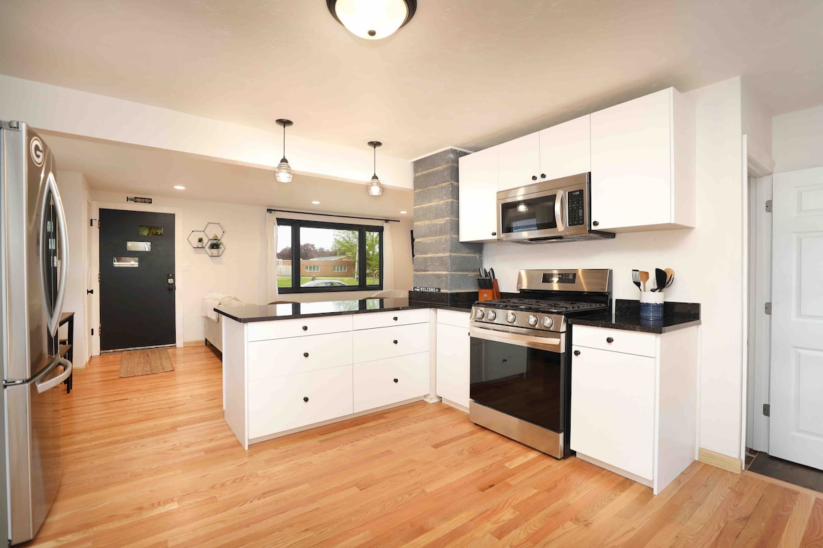 A modern kitchen features sleek white cabinetry and a black countertop. Stainless steel appliances, including a gas stove and microwave, are visible. Natural light enters through a large window, and a small seating area can be seen near the window.