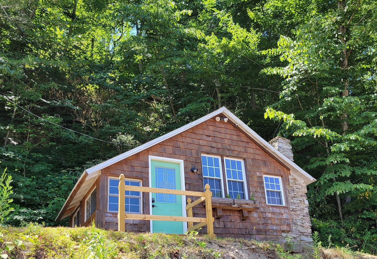 A rustic cabin is nestled among lush greenery, featuring a warm wood exterior with stone accents. The front porch, framed by a wooden railing, welcomes with a bright blue door and several windows allowing natural light.