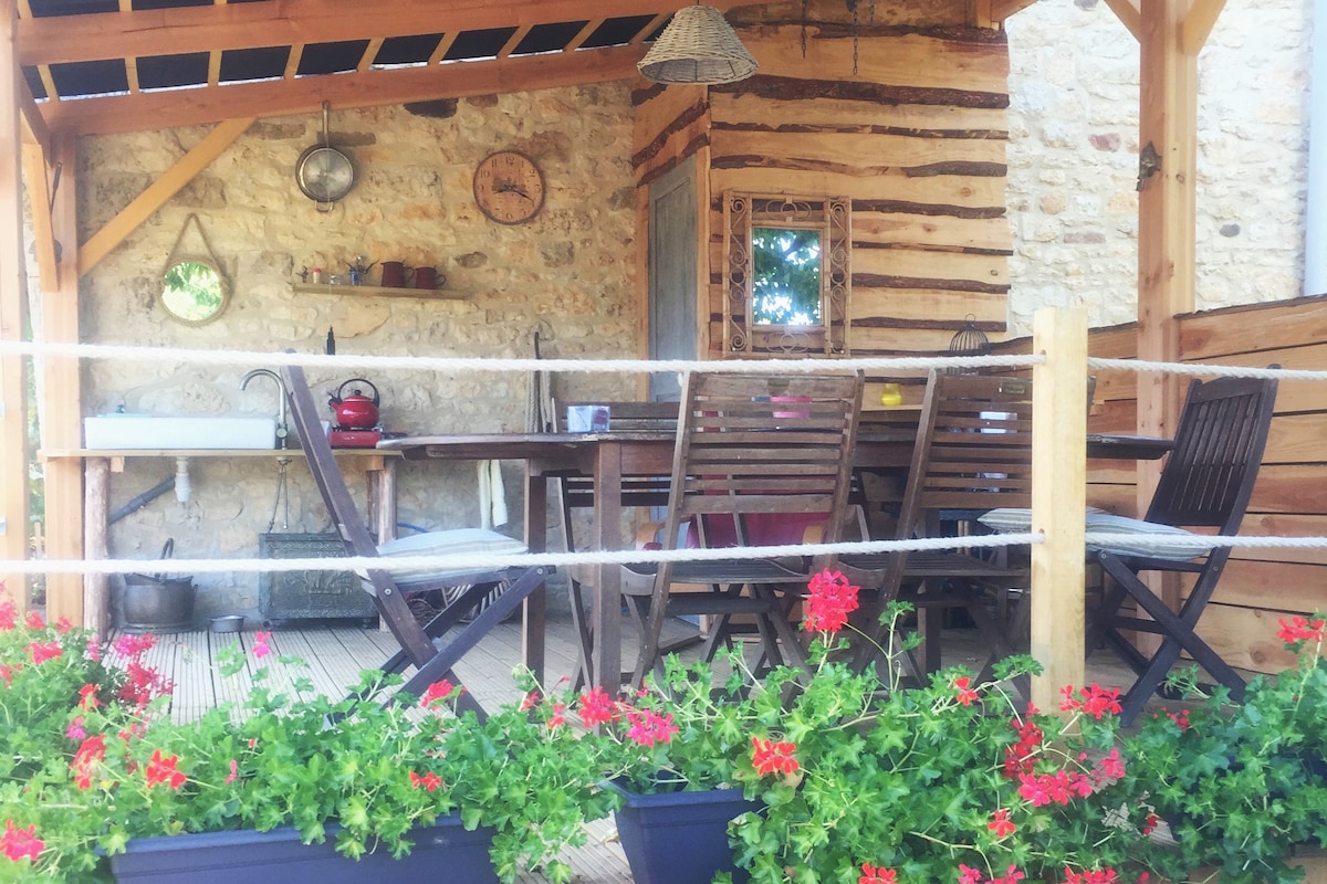 An outdoor dining area is surrounded by wooden beams and adorned with vibrant red geranium flowers. A large table with several chairs invites gatherings, while a simple sink and wall clock add functional charm to the rustic setting.